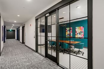 A hallway with a carpeted floor, a table with chairs, and a glass wall with artwork on it.